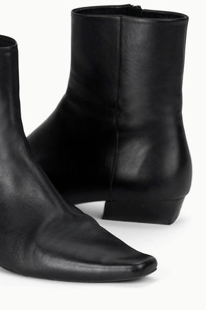Wally Flat Ankle Boot in Black displayed on a rustic surface, emphasizing its trendy design and contrasting materials.